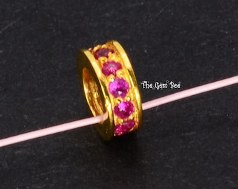 May include: A gold-toned cylindrical bead featuring a central band of vibrant, round-cut pink gemstones. The bead is threaded onto a thin, light pink string. The text The Gem Bee is visible in white lettering to the right of the bead.