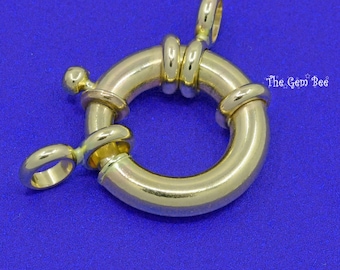May include: A polished gold-tone circular spring ring clasp featuring a smooth, rounded metal band with a small spring-loaded lever and an attached jump ring. The text The Gem Bee is visible in the upper right corner of the image.