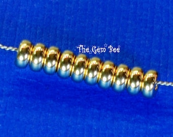 May include: A row of ten shiny, metallic gold-toned round beads strung on a thin silver wire. The beads have a smooth, reflective surface and are arranged horizontally. The text "The Gem Bee" is printed in white above the center of the beads.