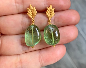 May include: A pair of gold-toned earrings featuring a leaf-shaped top component and a dangling, smooth, translucent green gemstone. The earrings have a polished finish and a vertical metal pin running through the center of each green stone.