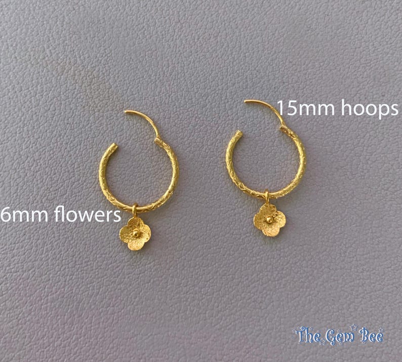 Puede incluir: Un par de pendientes de aro dorados con acabado texturizado, cada uno con un dije de flor de 6mm. Los aros miden 15mm de di&aacute;metro. El texto 15mm hoops y 6mm flowers es visible, junto con la marca The Gem Bee en la esquina inferior.