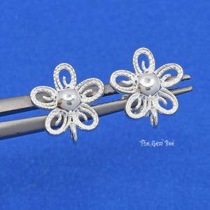 May include: Two silver-colored flower-shaped earring components. Each component has a central bead and five petals with a textured design. The components are held by a pair of tweezers against a blue background. The Gem Bee logo is in the bottom right corner.