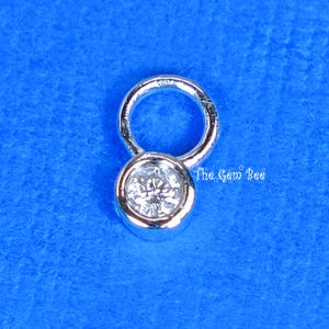 Peut inclure: Un pendentif en métal argenté avec une pierre ronde transparente à facettes sertie dans une monture en biseau. Le bijou comporte un anneau de jonction circulaire sur le dessus. Le texte The Gem Bee est imprimé en blanc près du pendentif.