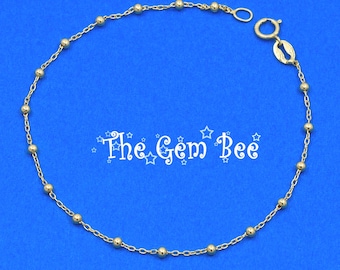 May include: A delicate gold chain bracelet featuring evenly spaced small gold beads along the length of the chain. The bracelet includes a circular spring ring clasp and a small oval tag. The text The Gem Bee is centered in the middle.