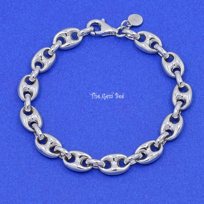 May include: A silver-toned, polished link bracelet with a lobster clasp closure. The links are oval-shaped with a rounded, hollowed-out center. The Gem Bee is printed on the blue background.