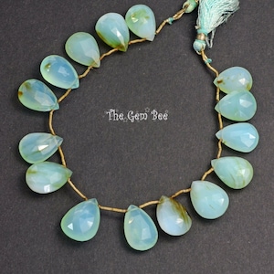 May include: A strand of light green and brown teardrop-shaped gemstone beads strung on a gold wire. The beads are faceted and have a translucent appearance. The strand is finished with a light blue tassel.