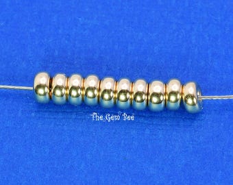 May include: A row of ten smooth, metallic gold-toned round beads strung on a thin silver wire. The beads have a reflective, polished surface and are arranged horizontally. The text The Gem Bee is printed in white below the center of the beads.