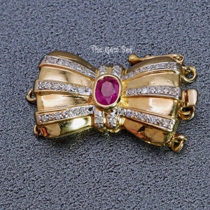 Könnte beinhalten: Goldener bogenförmiger Verschluss mit einem Rubinstein und Diamantbesatz. Der Verschluss ist für eine Halskette oder ein Armband gedacht.