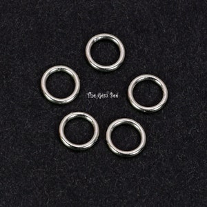 May include: Six silver jump rings, also known as split rings, are arranged in a circle on a black surface. The rings are all the same size and are made of metal.