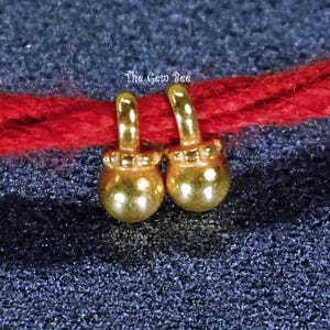 May include: Two small, shiny gold-toned charms with rounded, spherical bases and looped tops, suspended from a thick red string. The text The Gem Bee is printed in white above the charms. The charms feature a textured, beaded collar detail.