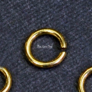 May include: Gold jump rings, a type of jewelry finding, are shown on a dark gray surface. The jump rings are round and have a smooth finish.