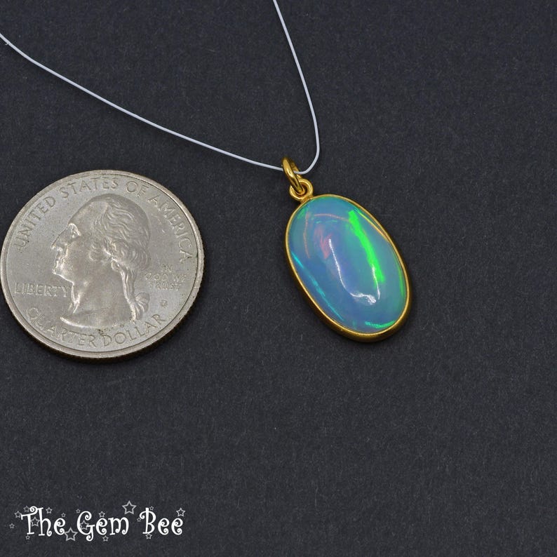 May include: An oval opal pendant with a gold setting, suspended on a white cord. The opal displays blue and green iridescence. The pendant is next to a quarter coin for size comparison. The gemstone is approximately 2 cm in length.