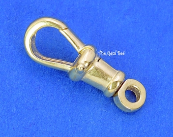 May include: A gold-toned metal lobster claw clasp featuring a spring-loaded gate and a swivel base with a circular attachment loop. The surface is polished and reflective. The text The Gem Bee is printed in white near the center of the clasp.