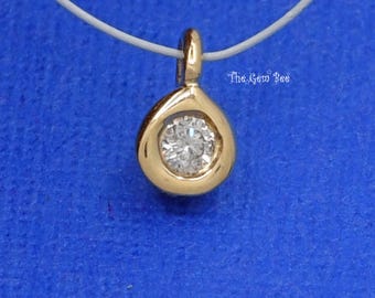 May include: A gold teardrop-shaped pendant featuring a single round brilliant-cut clear gemstone set in the center. The pendant includes a small bail at the top and is suspended by a thin white cord. The text The Gem Bee is visible.