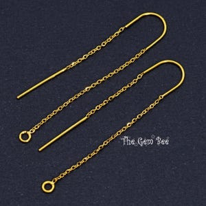 May include: A pair of gold-tone threader earrings featuring a long, slender curved wire connected to a delicate cable chain. Each chain ends in a small circular jump ring. The text The Gem Bee is visible in the lower right corner of the image.