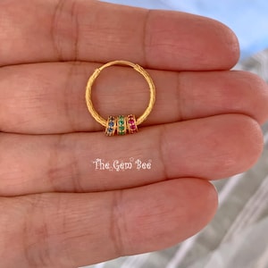 Puede incluir: Un pendiente de aro dorado con superficie texturizada y tres peque&ntilde;os anillos m&oacute;viles adornados con piedras preciosas azules, verdes y rojas. El texto The Gem Bee aparece debajo del aro. El dise&ntilde;o incluye un cierre de alambre fino.
