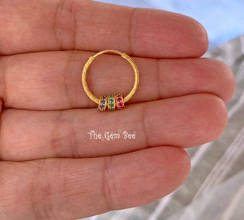 Puede incluir: Un pendiente de aro dorado con superficie texturizada y tres peque&ntilde;os anillos m&oacute;viles adornados con piedras preciosas azules, verdes y rojas. El texto The Gem Bee aparece debajo del aro. El dise&ntilde;o incluye un cierre de alambre fino.