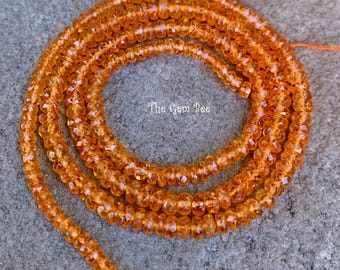 May include: A long strand of faceted, translucent orange gemstone beads arranged in a circular coil. The beads are uniform in size and shape, featuring a reflective surface. The text The Gem Bee is printed in white in the center of the coil.