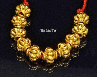 May include: A necklace featuring nine gold-toned, ribbed, barrel-shaped beads strung on a thin red cord. The beads have a metallic finish and a repeating vertical grooved pattern. The text "The Gem Bee" is printed in white in the center of the arc.