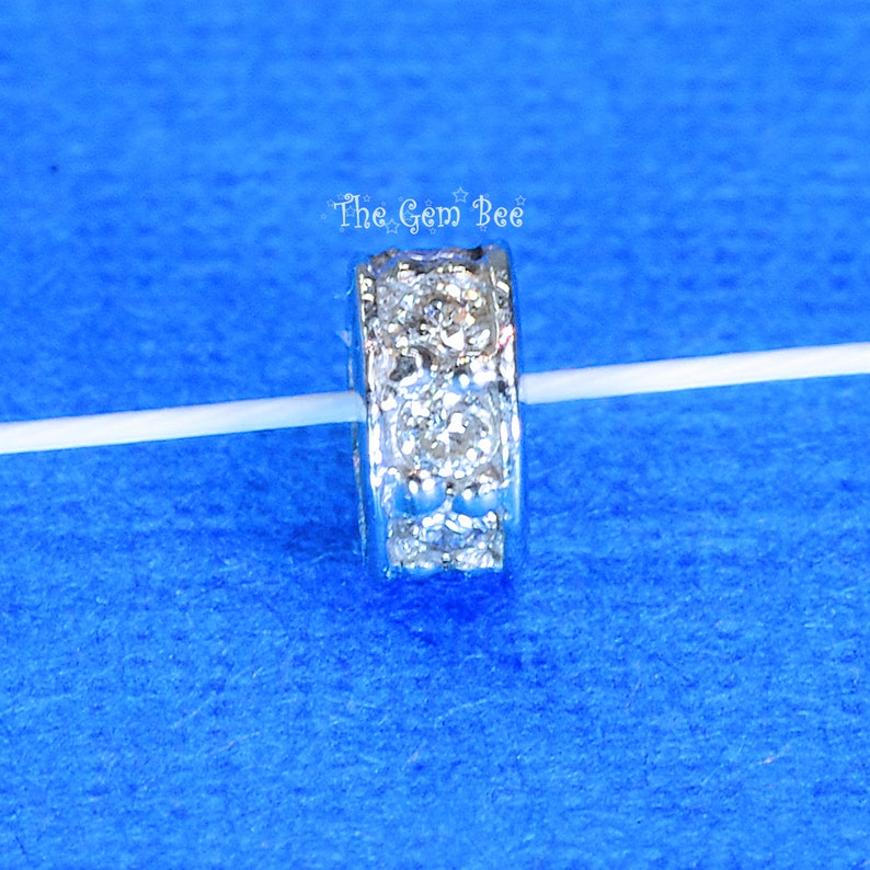 May include: Silver spacer bead with a row of clear cubic zirconia stones around the edge. The bead is on a white string.