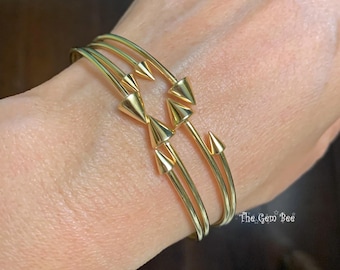 May include: A gold-toned cuff bracelet featuring two thin, parallel bands that meet at a central point. The design includes six small, polished cone-shaped spikes arranged in a symmetrical pattern. The text The Gem Bee is visible in the corner.