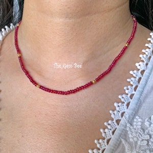 Może przedstawiać: Delikatny naszyjnik z czerwonych kamieni szlachetnych z złotymi koralikami. Naszyjnik znajduje się na szyi modelki i ma około 40 cm długości.