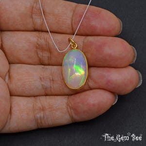 May include: Oval opal pendant with a gold-coloured bezel and bail, suspended on a white cord. The opal displays iridescent colours of green, yellow, and blue. The Gem Bee logo is visible in the bottom right corner.