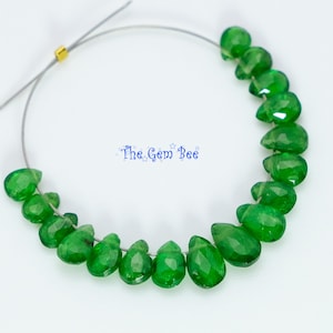 May include: A strand of faceted, teardrop-shaped green gemstones arranged in a curved line on a thin wire. A small gold-colored metal bead is visible on the wire. The text "The Gem Bee" is printed in blue in the center of the image.