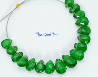 May include: A strand of faceted, teardrop-shaped green gemstones arranged in a curved line on a thin wire. A small gold-colored metal bead is visible on the wire. The text "The Gem Bee" is printed in blue in the center of the image.