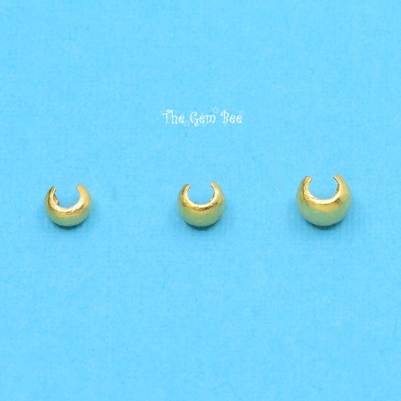 May include: Three gold-coloured metal crimp beads, each with a rounded shape and a small opening. The beads are arranged in a row on a blue background.