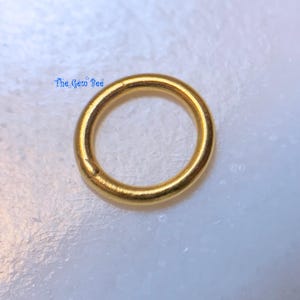 May include: A single circular gold jump ring with a smooth, polished finish. The text The Gem Bee is printed in blue near the upper left side of the ring. The metal ring has a consistent thickness and a closed loop design for jewelry making.