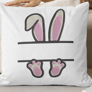 May include: White throw pillow with embroidered bunny ears and feet. The bunny ears are pink and the feet are pink with black outlines. The pillow has a blank space for personalisation.