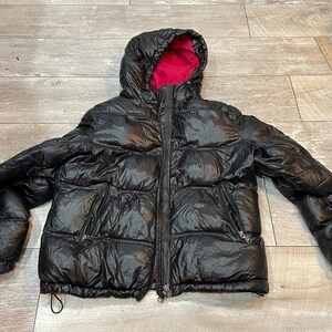 Peut inclure: Veste matelassée noire avec capuche et doublure intérieure rose vif. La veste a une fermeture éclair complète, deux poches latérales et un cordon de serrage à l'ourlet inférieur. Un manteau d'hiver.