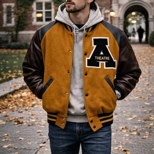 May include: A mustard yellow varsity jacket with dark brown leather sleeves, a black and white letter A patch with the word THEATRE, a gray hooded sweatshirt underneath, and blue denim jeans. The jacket features black and yellow striped ribbed trim.