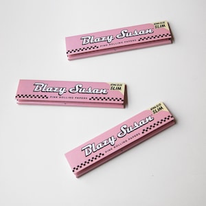 May include: Three pink Blazy Susan King Size Slim rolling paper packs. Each pack has the brand name in white script, the words "Pink Rolling Papers", and a black and white checkered pattern.