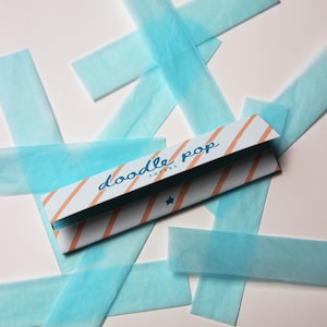 May include: A package of "doodle pop PAPERS" with orange diagonal stripes and a blue star, surrounded by light blue tissue paper strips. The text "doodle pop PAPERS" is written in a cursive font.