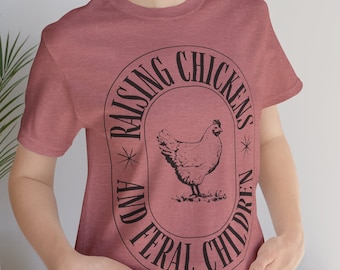 Könnte beinhalten: Ein heidefarbenes T-Shirt mit einem schwarzen Grafikdesign eines Huhns in einem Kreis. Der Text um den Kreis lautet "Raising Chickens and Feral Children".