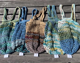 May include: Five handmade crochet market bags in various colors, including blue, green, and tan. Each bag has a unique color pattern and a sturdy handle. The bags are numbered 1 through 5 and are displayed on a wooden surface.