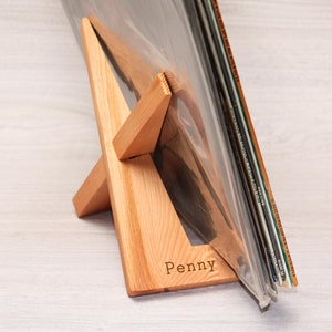 May include: A wooden record stand with a triangular base and a name engraved on the front. The stand is holding a stack of vinyl records.