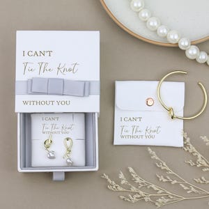 Puede incluir: Una caja de regalo blanca con un lazo morado claro y el texto "No puedo atar el nudo sin ti". La caja contiene un par de pendientes de oro con una perla y un diseño de nudo. Se muestra una segunda caja blanca con el mismo texto con una pulsera de oro con un diseño de nudo.