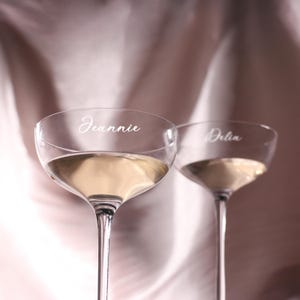Puede incluir: Dos copas de champ&aacute;n transparentes llenas de un l&iacute;quido claro. La palabra "Jeannie" est&aacute; escrita en una copa y "Delia" en la otra. Las copas tienen tallos largos y cuencos anchos y poco profundos.