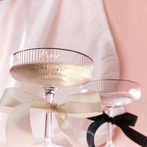 Puede incluir: Dos copas de champán transparentes con un diseño acanalado y borde dorado. Una copa tiene el nombre "Piper" impreso. Ambas copas tienen una cinta atada alrededor del tallo. Una cinta es de color crema y la otra es negra.