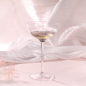 Puede incluir: Una copa de champ&aacute;n transparente con un tallo largo, llena de un l&iacute;quido de color claro. La palabra "Lucy" est&aacute; grabada en el cristal. La copa se encuentra sobre un fondo rosa suave con tela drapeada.