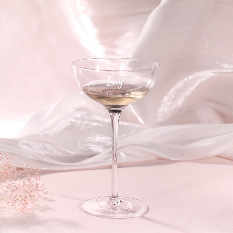Puede incluir: Una copa de champ&aacute;n transparente con un tallo largo, llena de un l&iacute;quido de color claro. La palabra "Lucy" est&aacute; grabada en el cristal. La copa se encuentra sobre un fondo rosa suave con tela drapeada.