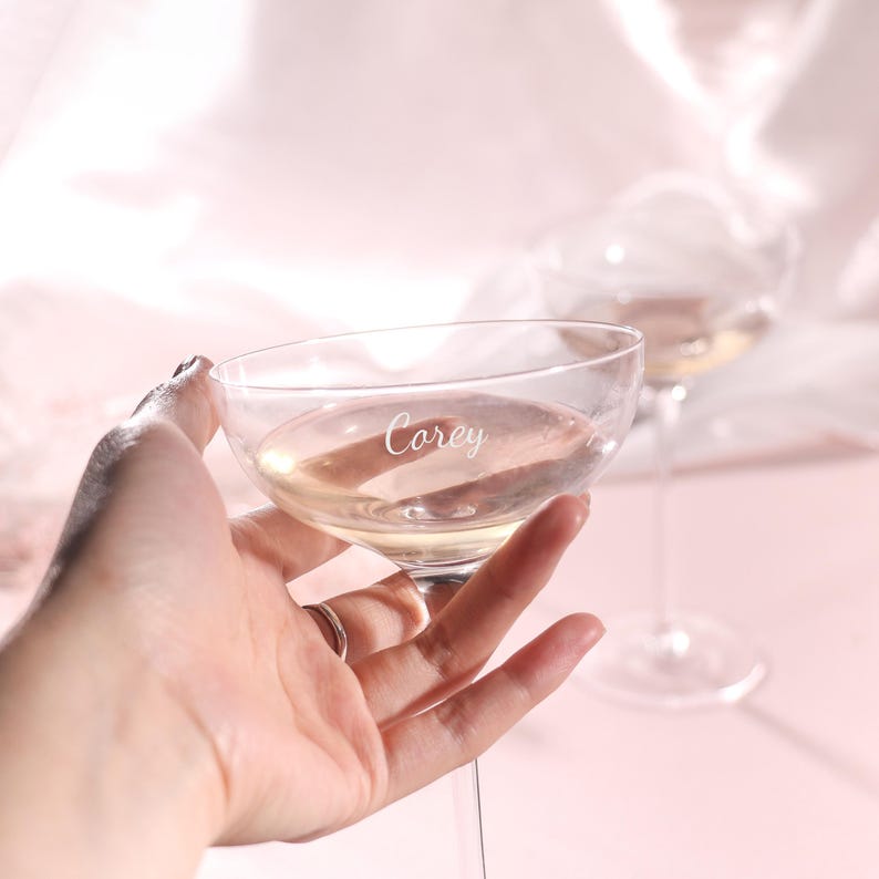 Puede incluir: Una copa de champ&aacute;n transparente llena de una bebida clara, sostenida por una mano. La palabra "Corey" est&aacute; grabada en el cristal. Otra copa es visible en el fondo, con un fondo rosa suave.