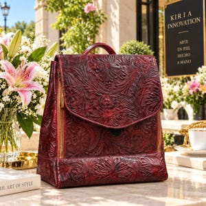 Puede incluir: Una mochila de cuero rojo oscuro con un intrincado patrón floral en relieve en toda la superficie, un asa superior y cremalleras verticales de tono dorado en los laterales. El bolso tiene un cierre de solapa estructurado y una base rectangular.