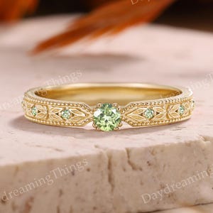 May include: A gold ring with a central light green gemstone and smaller green stones. The band has an ornate, engraved design. The ring is set against a neutral background with blurred orange and brown elements.