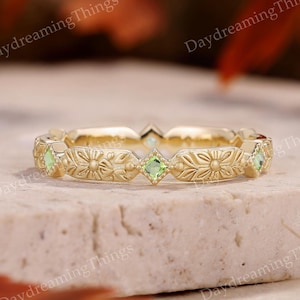 May include: A gold-colored ring with a floral design and green gemstones. The ring features alternating floral patterns and square-cut green stones. The ring is set on a neutral surface with a blurred background.
