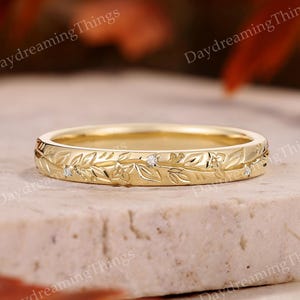 May include: A gold-colored ring with a floral and leaf pattern. The ring has small, clear gemstones set into the design. The ring is on a light-colored surface with the text "Daydreaming Things" in the background.