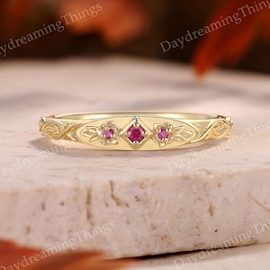Könnte beinhalten: Ein goldener Bandring mit drei kleinen, runden, leuchtend rosa Edelsteinen in einer zentralen rautenförmigen Fassung, flankiert von filigranen, eingravierten Blumenblattmustern entlang der Ringschiene, mit dem Text DaydreamingThings.